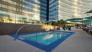 Una piscina interior moderna con agua azul cristalina, rodeada de sillones de descanso y sombrillas, y un edificio con fachada de vidrio al fondo.
