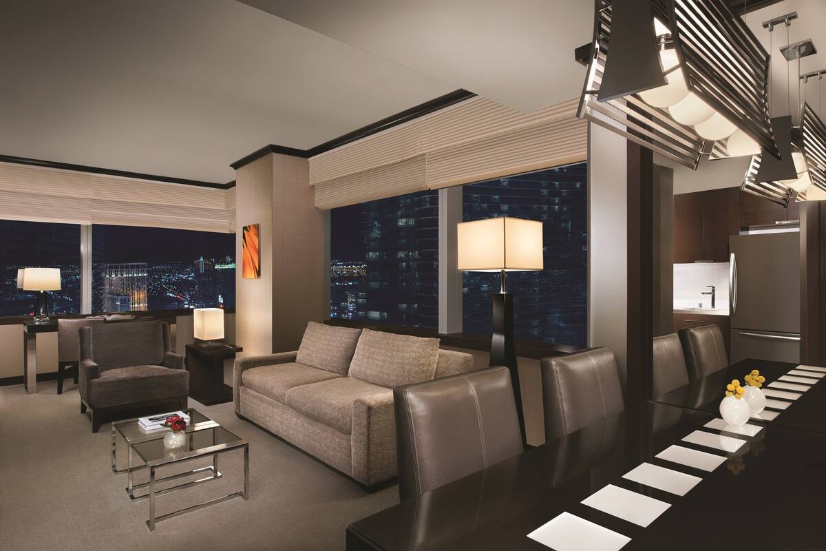 Una habitación de hotel moderna con vista a la ciudad, un sofá, una mesa de comedor con vajilla y un ventanal grande con persianas.