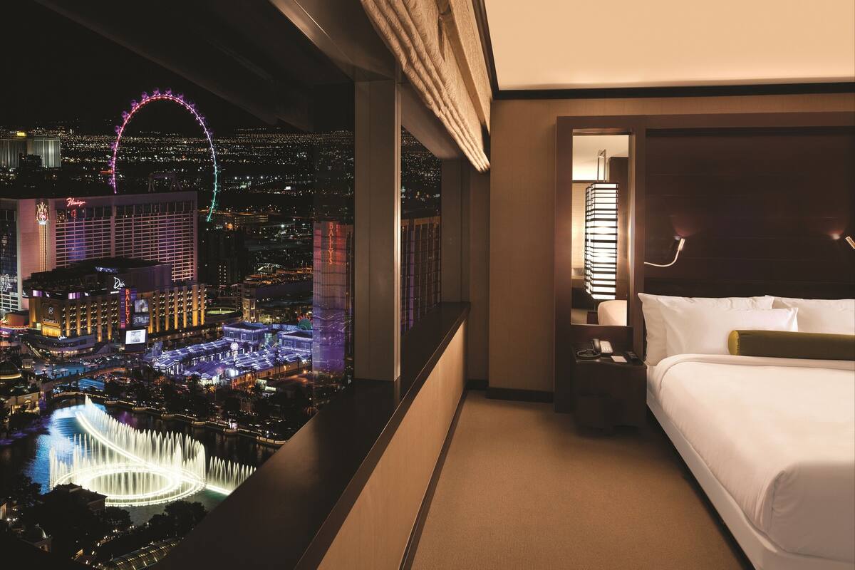 Habitación de hotel con cama, mesita de noche y vista a un paisaje urbano que incluye una noria.