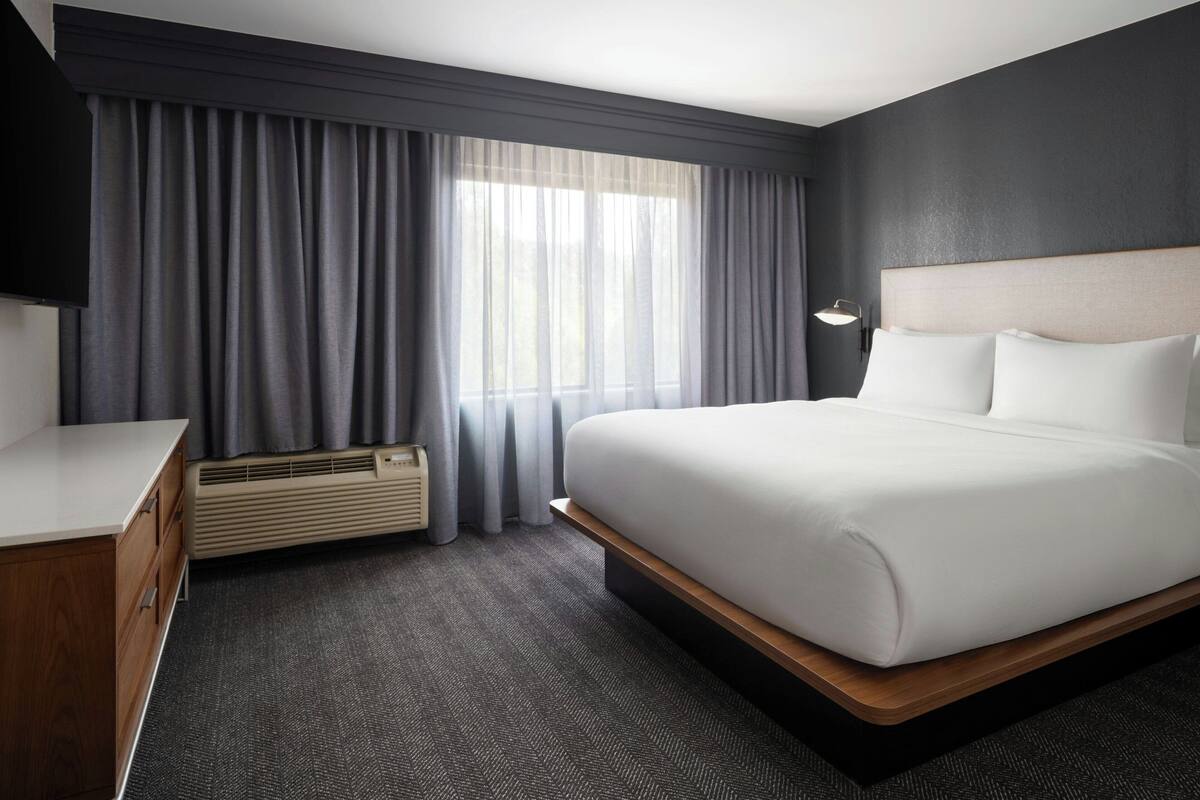 Ein modernes Hotelzimmer mit einem großen Bett, einem Nachttisch, einem Flachbildfernseher und einem Fenster mit Vorhängen.