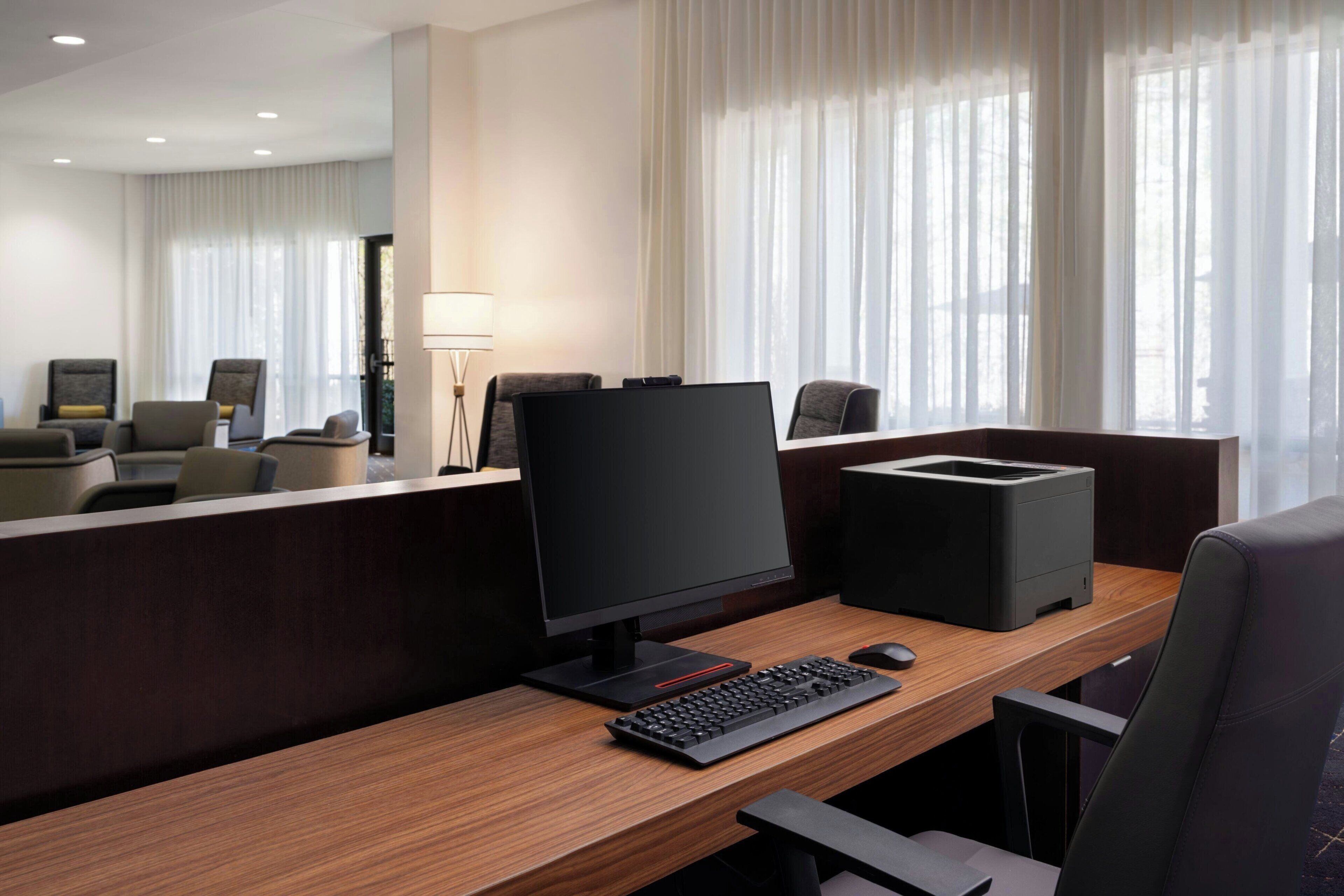 A hotel room with a wooden desk, a computer, a printer, and a chair.