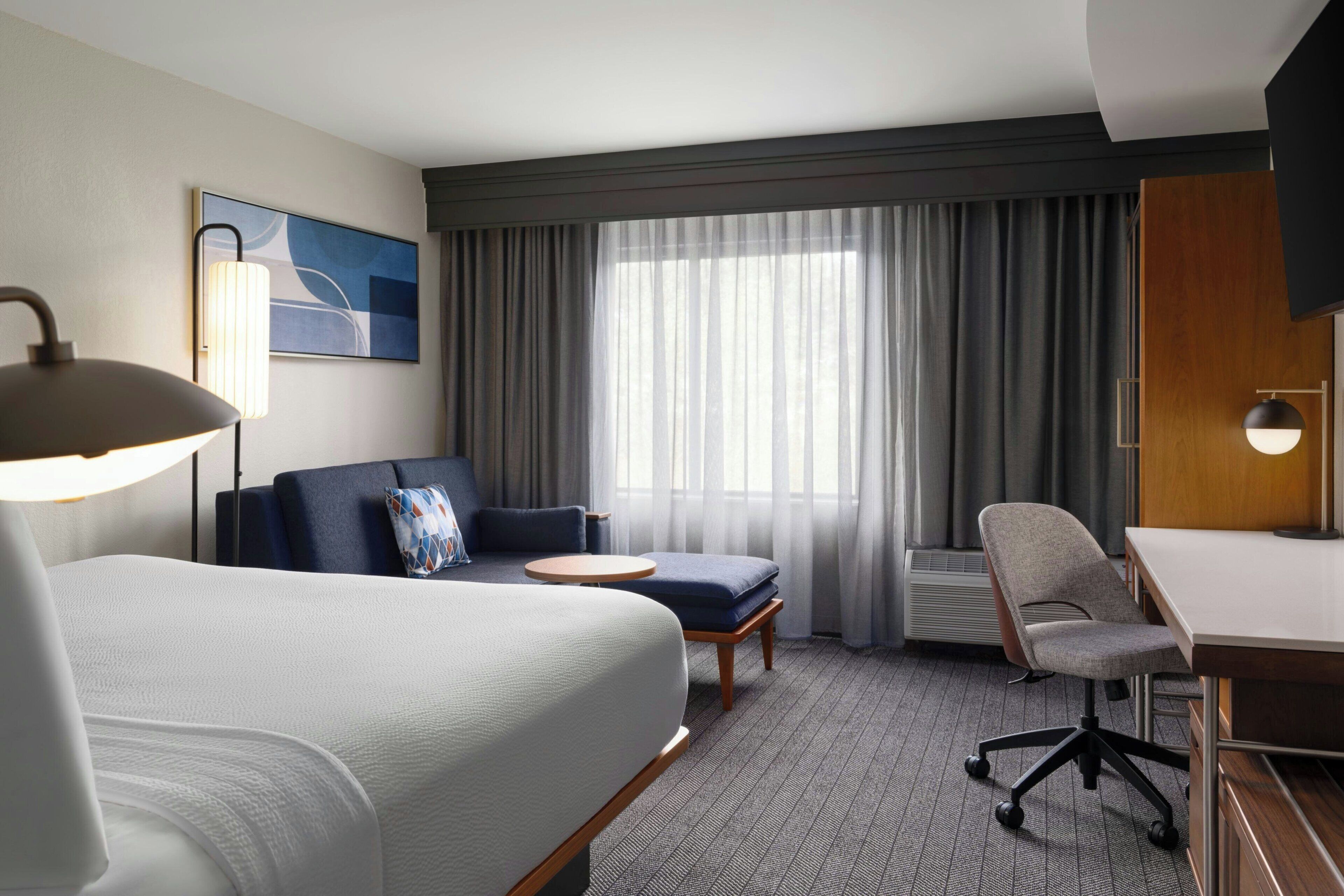 A hotel room with a bed, a desk, a chair, a sofa, a small table, a lamp, a painting, and a large window with curtains.