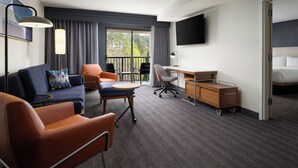 Hotelkamer met een bank, stoelen, een bureau, een televisie en een balkon met gordijnen.