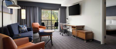 Une chambre d’hôtel avec un canapé, des fauteuils, un bureau, une télévision et un balcon avec des rideaux.