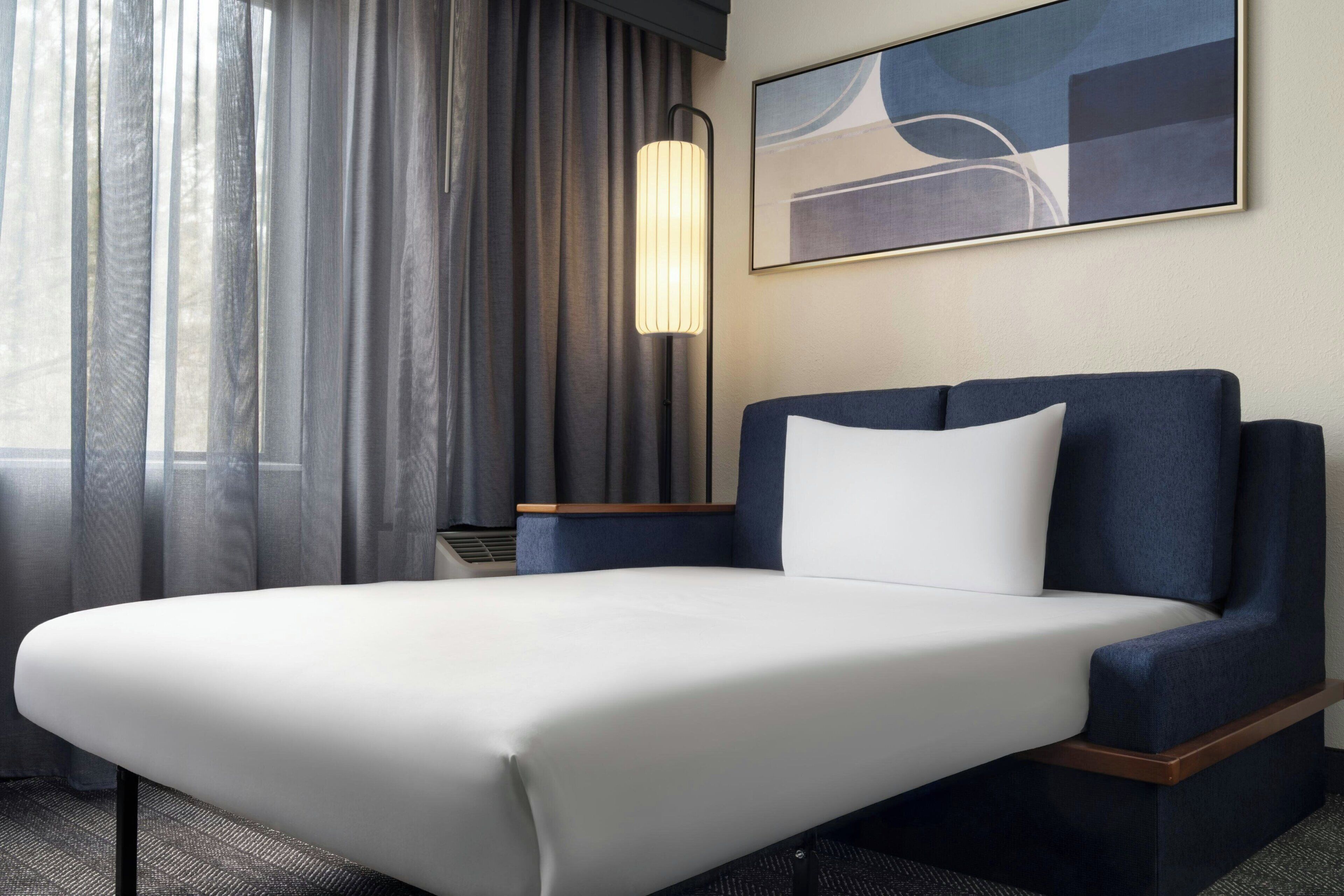 Quarto de hotel com cama, mesa de cabeceira, luminária, pintura e janela com cortinas.