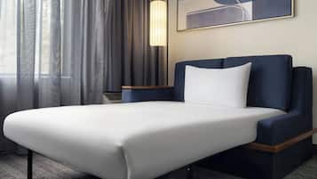 Una habitación de hotel con cama, mesita de noche, lámpara, un cuadro y ventana con cortinas.