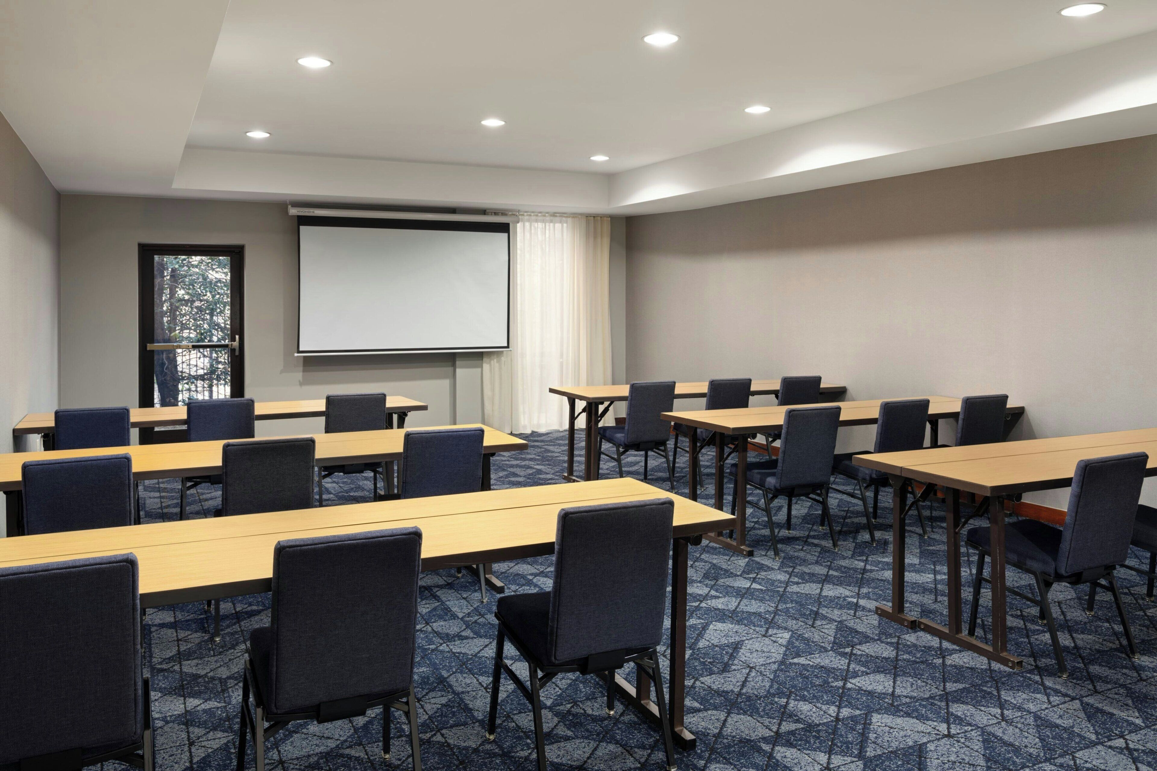 A conference room with multiple tables and chairs, a projector screen, and a large window with a view of trees.