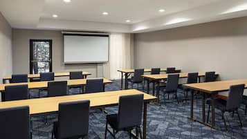A conference room with multiple tables and chairs, a projector screen, and a large window with a view of trees.