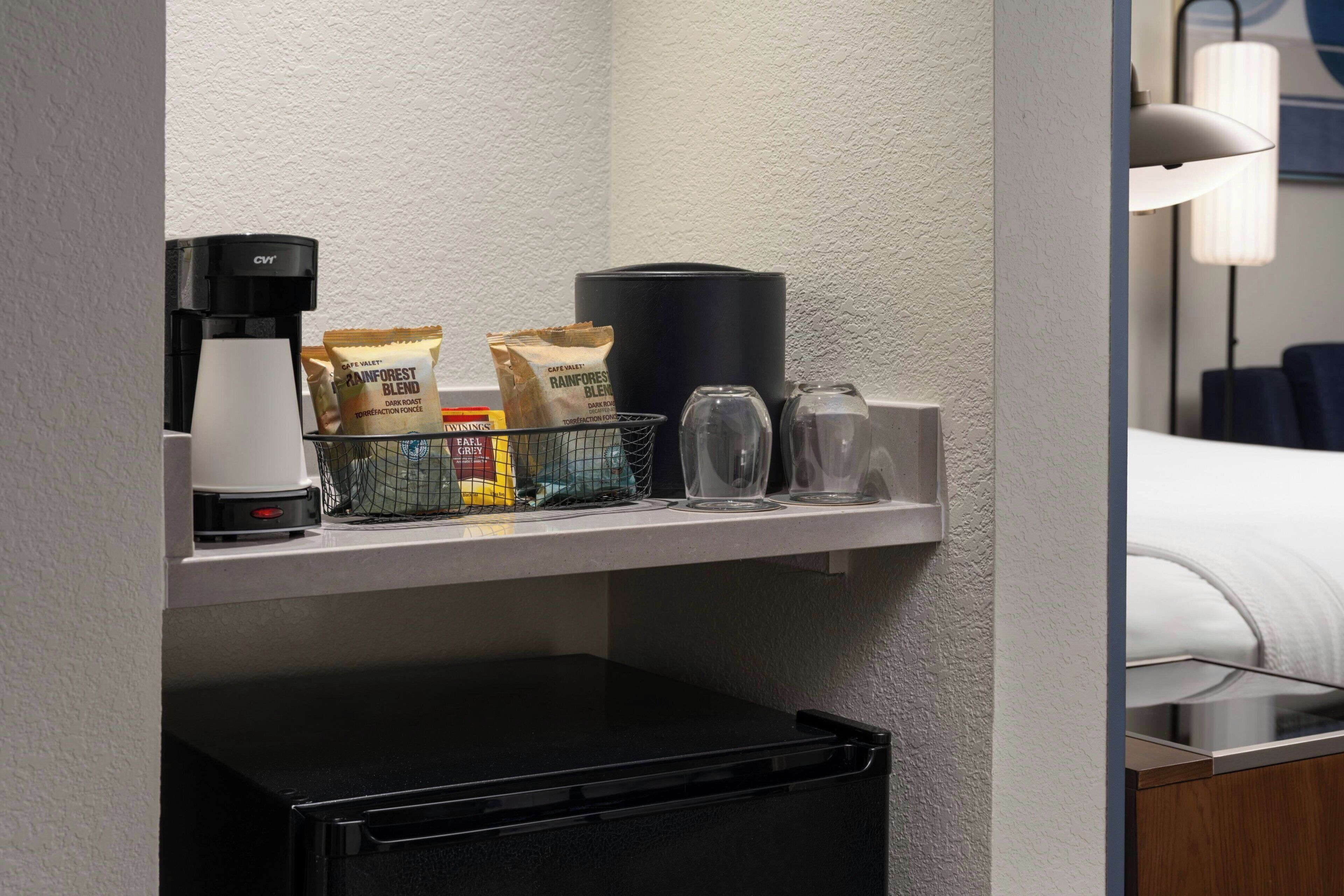 A hotel room with a minibar, a coffee maker, a lamp, and a bed.