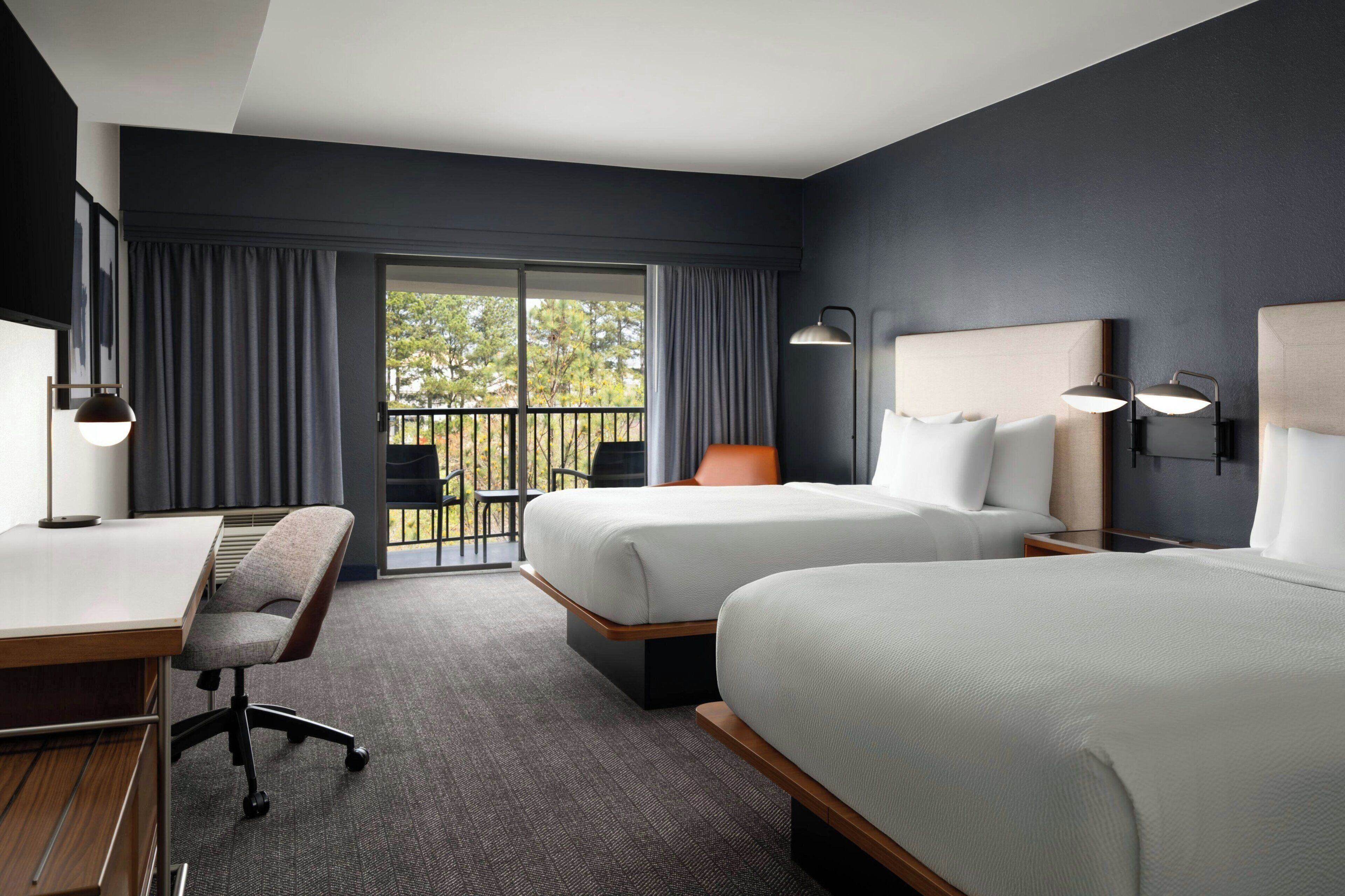 A hotel room with two beds, a desk, a chair, and a balcony with a view of trees.