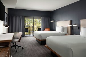 Habitación de hotel con dos camas, un escritorio, una silla y un balcón con vistas a árboles.