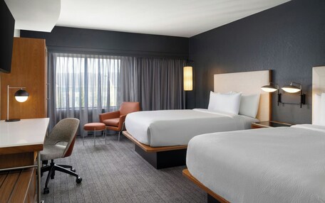 A hotel room with two beds, a desk, a chair, and a large window with curtains.