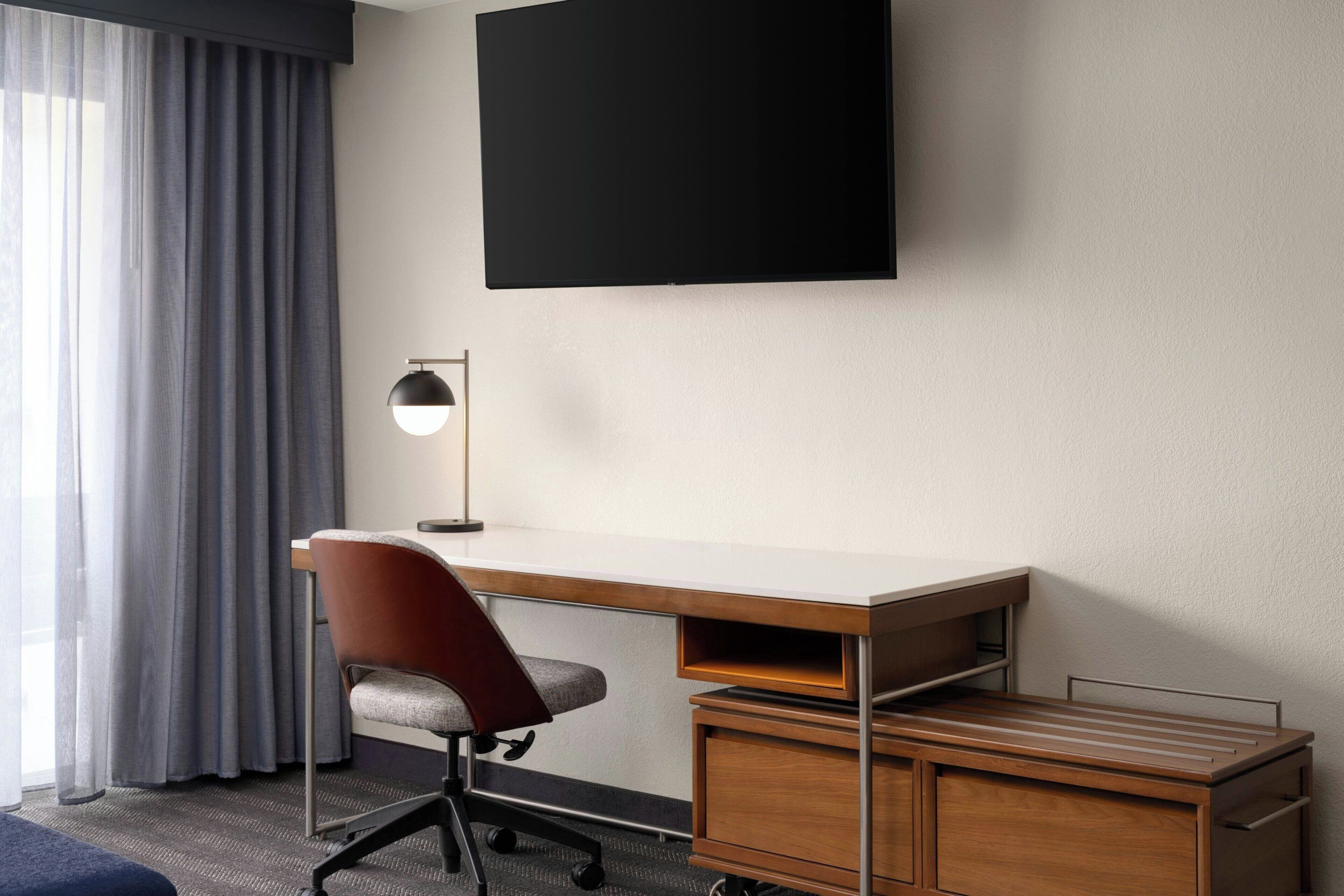 A hotel room with a desk, chair, TV, and a lamp.