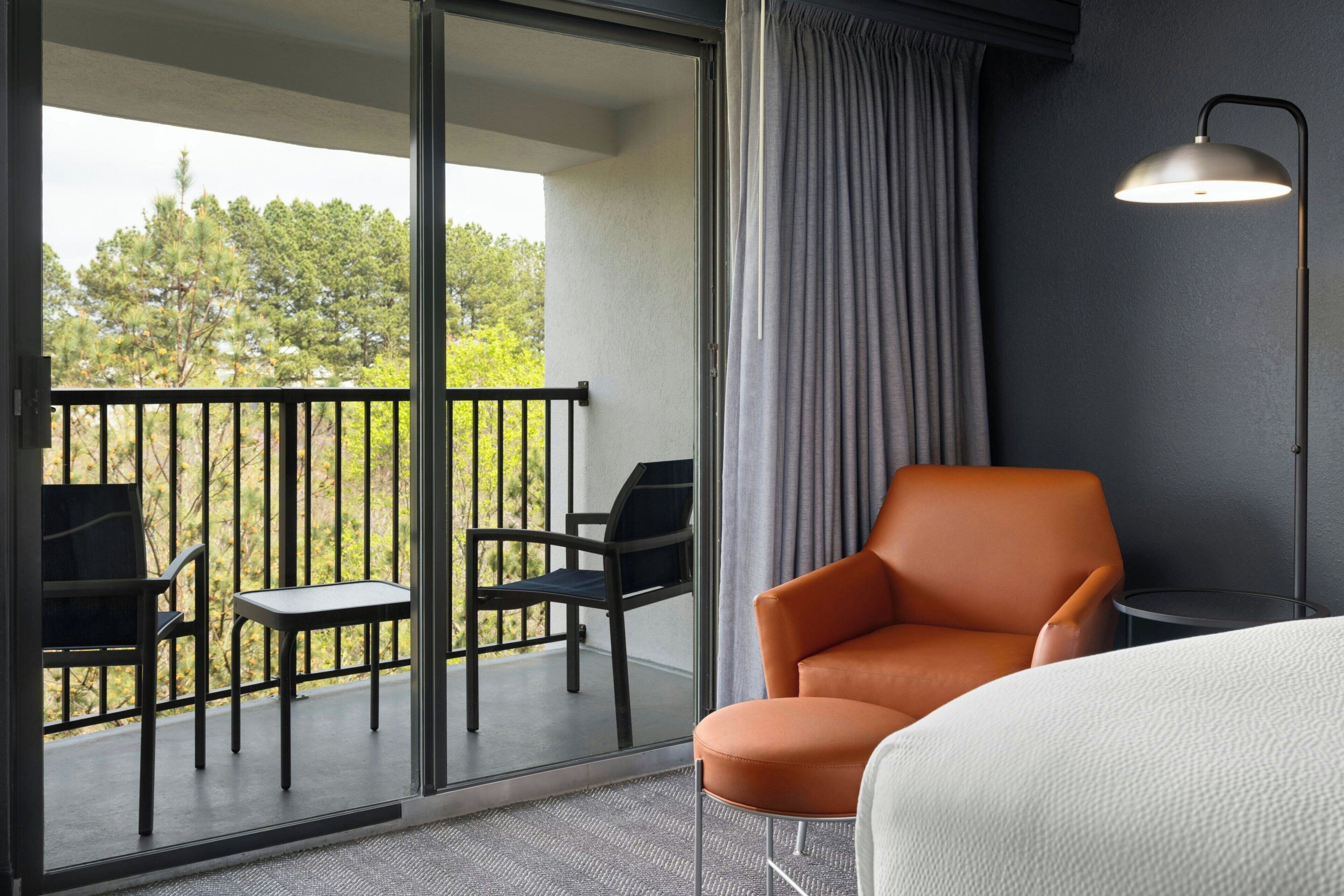 A hotel room with a balcony, a single orange chair, a small round table, a bed, and a floor lamp.