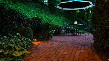 A brick pathway leading to a table and chairs with an umbrella, surrounded by lush greenery.
