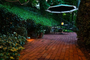 A brick pathway leading to a table and chairs with an umbrella, surrounded by lush greenery.