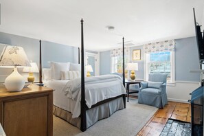 Una cama con dosel y tablero de madera, una mesita de noche con lámpara, una silla y vistas al exterior por una ventana.