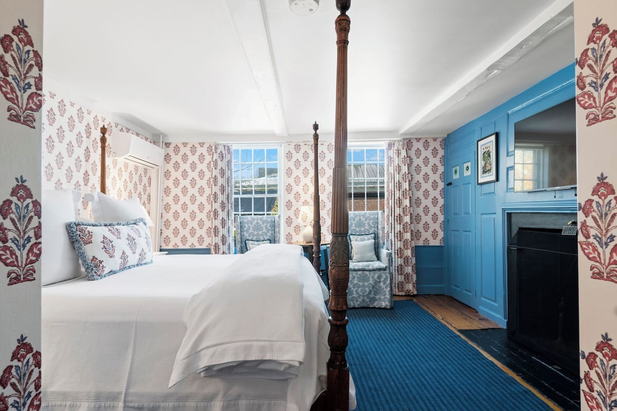 A bedroom with a four-poster bed, a blue armchair, a small table, and a fireplace.