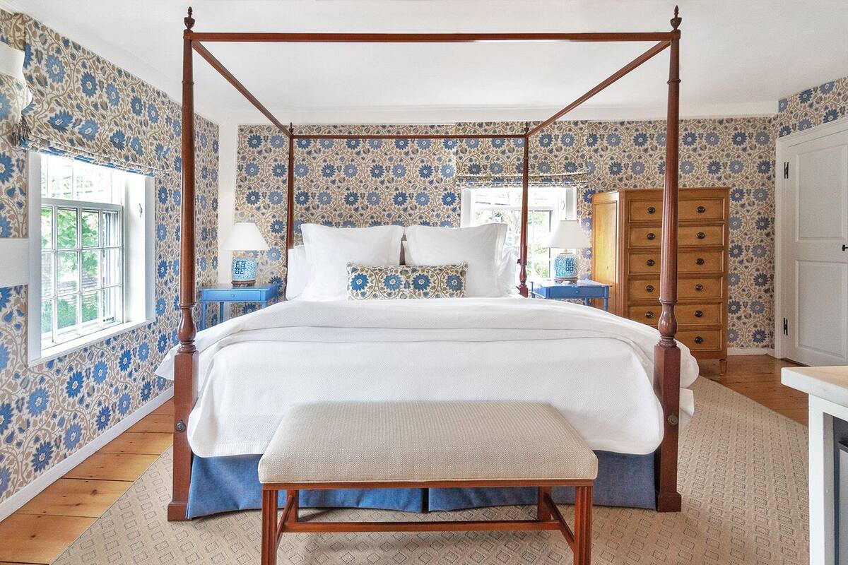 A four-poster bed with a canopy, a wooden dresser, and a bench in a room with floral wallpaper.
