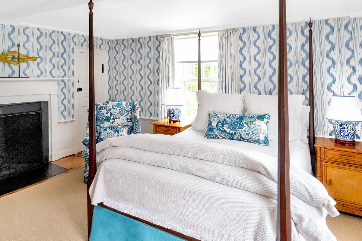 A four-poster bed with a white and blue patterned bedspread, two matching pillows, a bedside table with a lamp, and a fireplace with a blue and white patterned wallpaper.