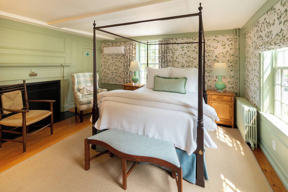 A four-poster bed with a bench, a fireplace, and a window with floral wallpaper.