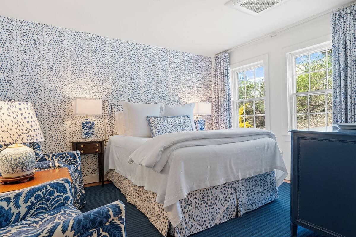 A bedroom with a patterned wallpaper, a bed with white bedding, a blue armchair, a nightstand with a lamp, and a dresser.