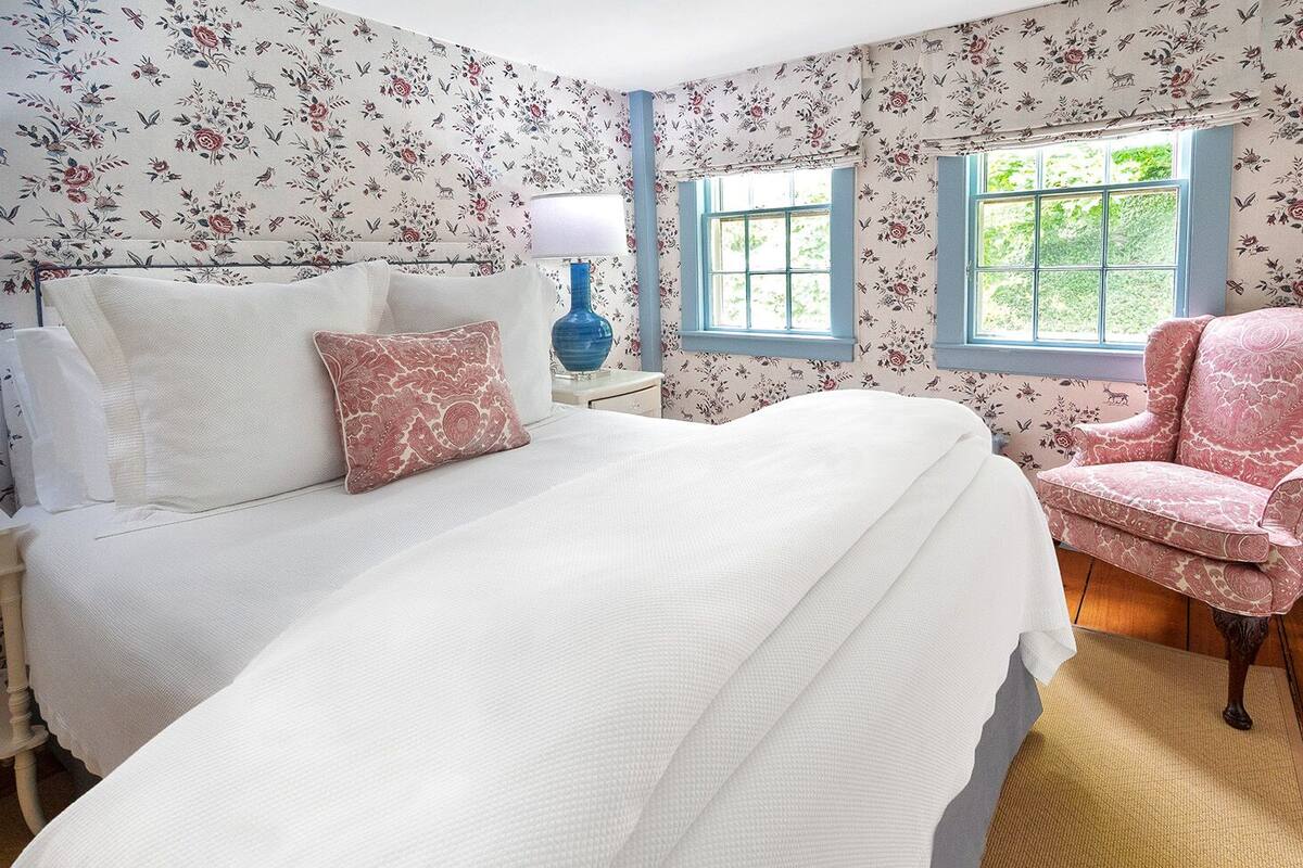A bedroom with floral wallpaper, a bed with white bedding, a blue lamp, and a pink armchair.