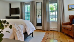 A bedroom with a bed, a TV, a window with curtains, a chair, and a painting on the wall. - Union Street Inn (Nantucket)