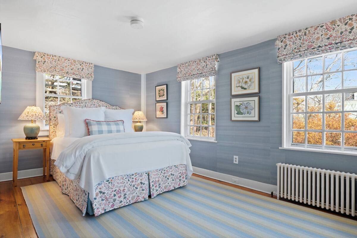A bedroom with a bed, two bedside tables, a window with floral valance, and a radiator.