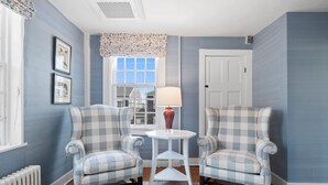 A room with two checkered armchairs, a small white table with a lamp, a window with a view of a building, and a radiator. - Union Street Inn (Nantucket)
