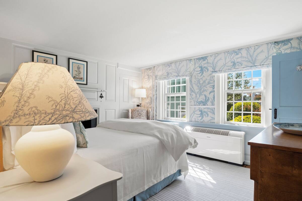 A bedroom with a bed, a bedside table with a lamp, a window with a view of greenery, and a radiator.