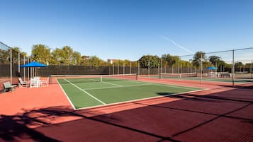 An outdoor tennis court with a net, seating area, and two blue umbrellas.