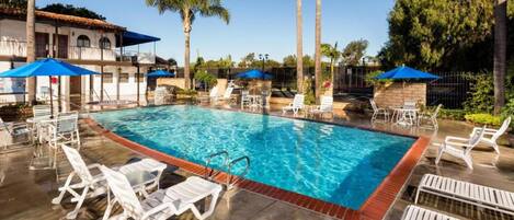 A swimming pool with lounge chairs and umbrellas, surrounded by palm trees and a building with a tiled roof.