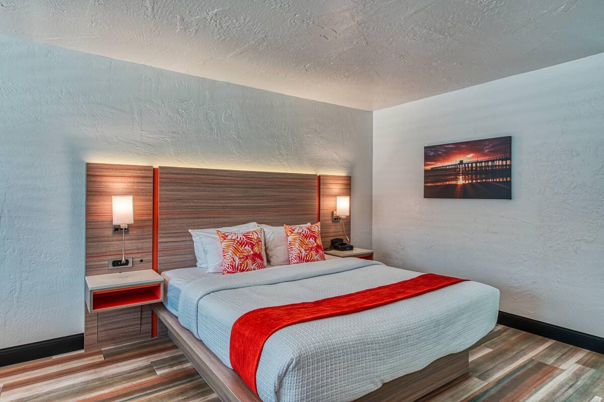 Une chambre d’hôtel avec un lit, des tables de chevet, un plaid rouge, une lampe fixée au mur et un tableau représentant un coucher de soleil sur une jetée.