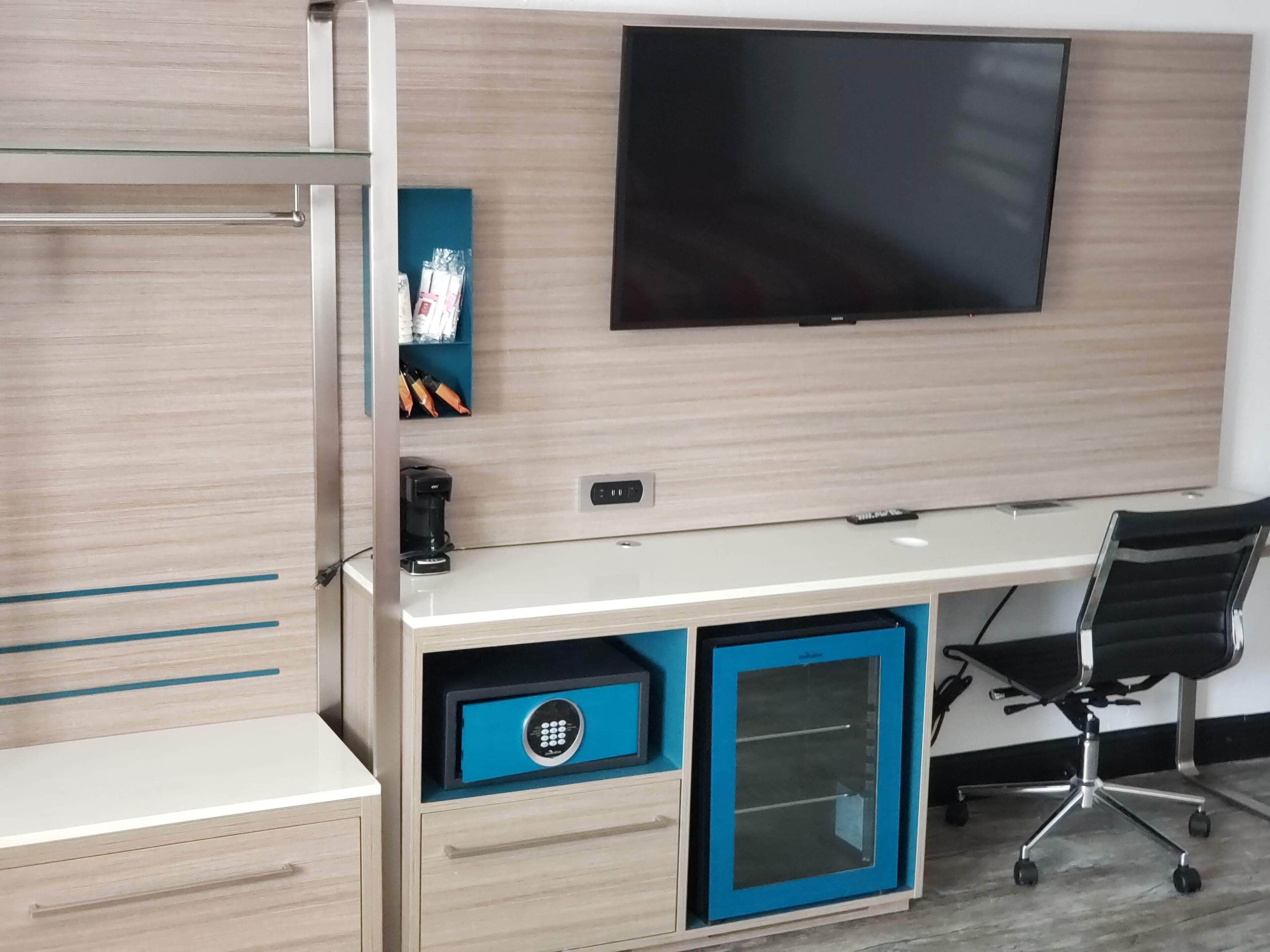 A modern office space with a large flat-screen TV mounted on the wall, a white desk with a blue drawer, and a black office chair.