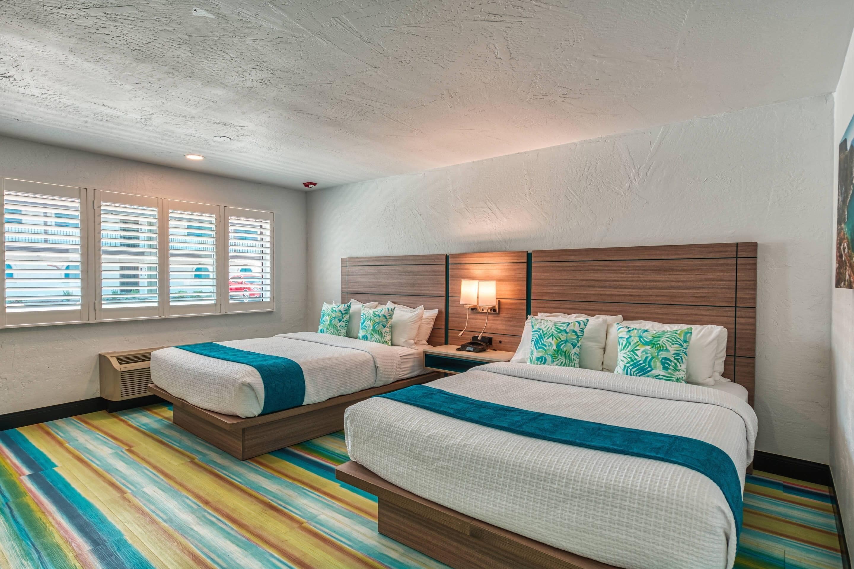 Habitación de hotel con dos camas, cabecero de madera, mesita de noche con lámpara y alfombra a rayas de colores.