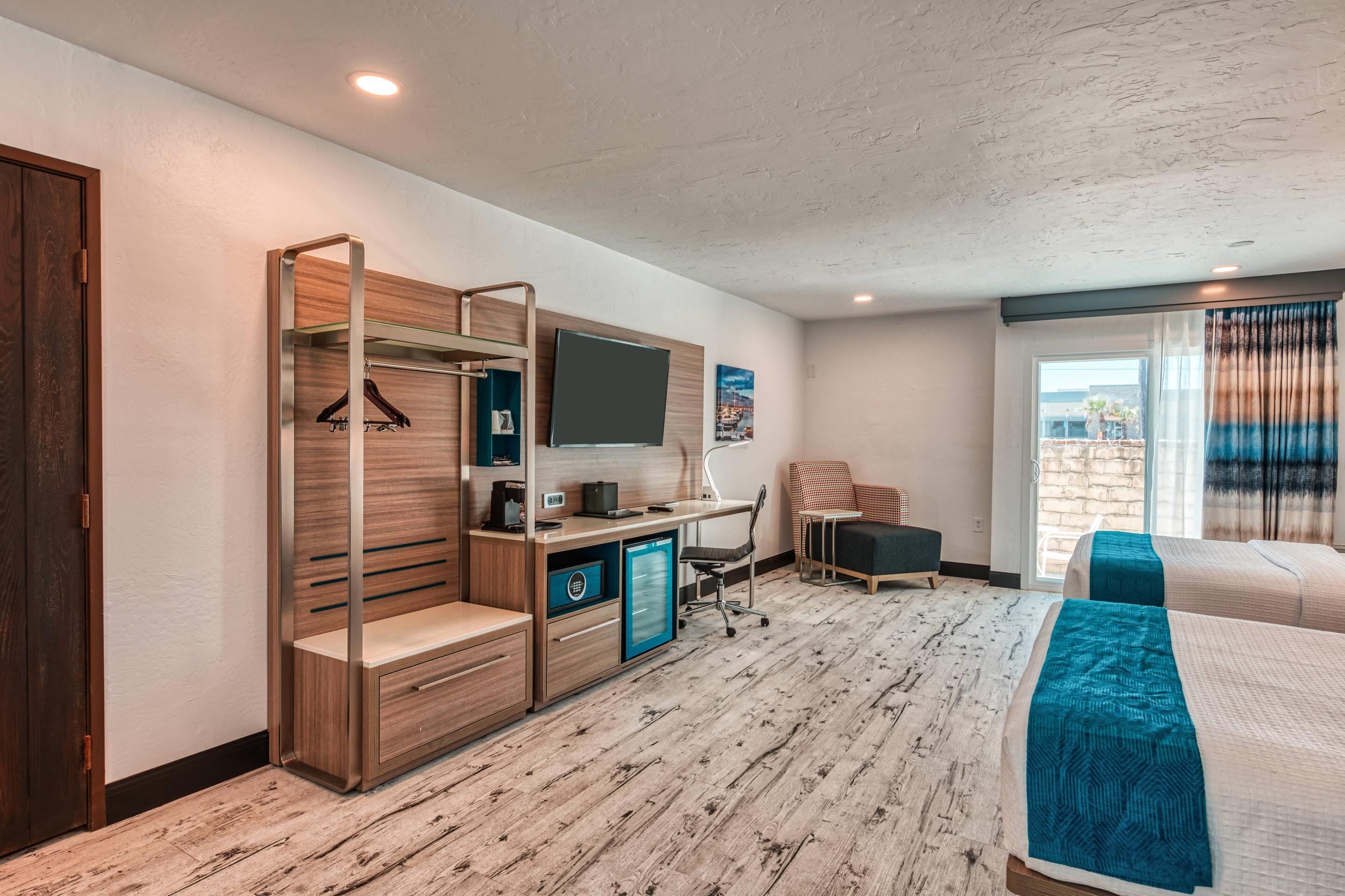 A hotel room with a wooden desk, a TV, a bed with a blue blanket, and a view of the outdoors through a window.