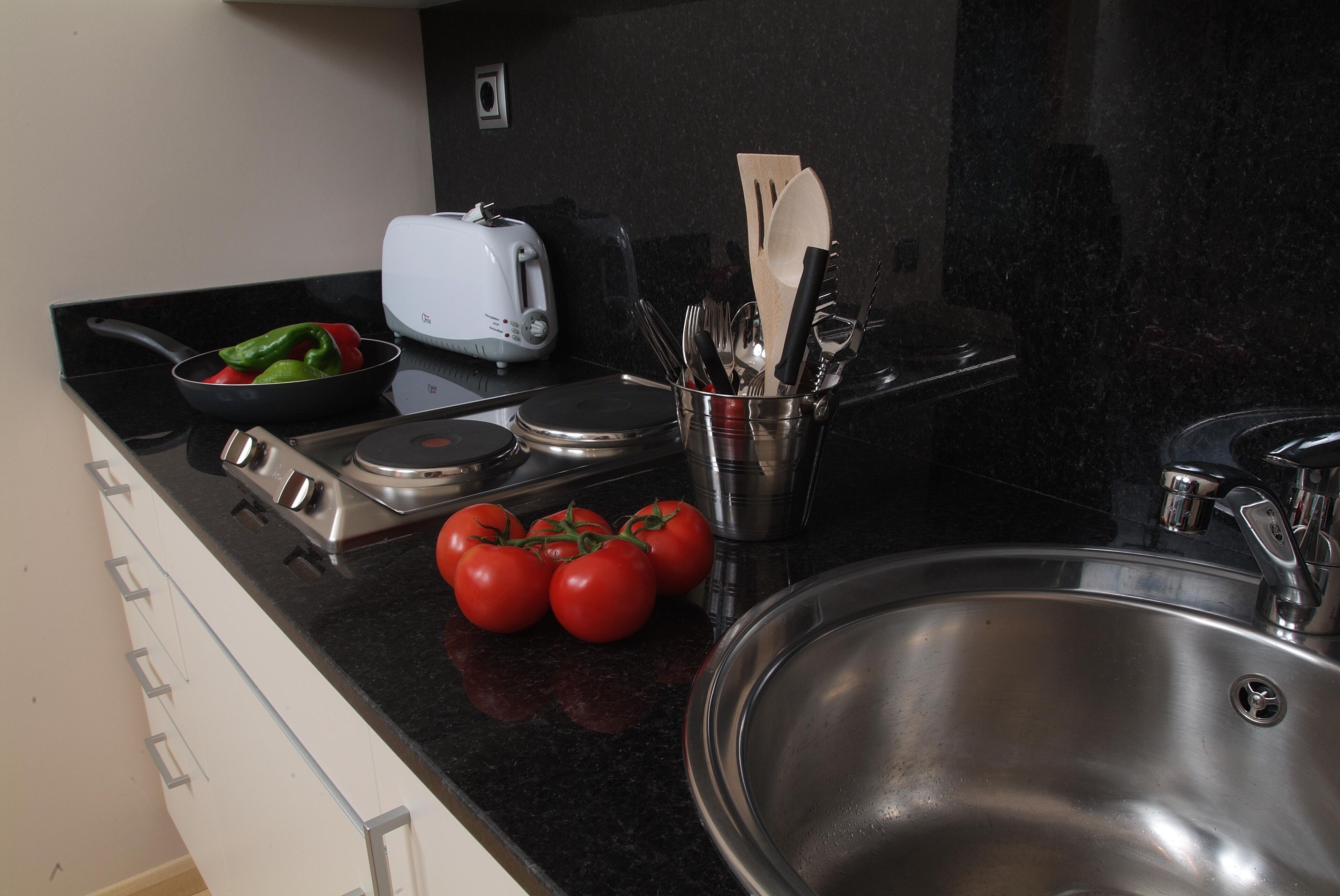 A kitchen with a black countertop, a stainless steel sink, a toaster, a frying pan, a pot, a saucepan, a wooden spatula, a metal utensil holder, and a few tomatoes.