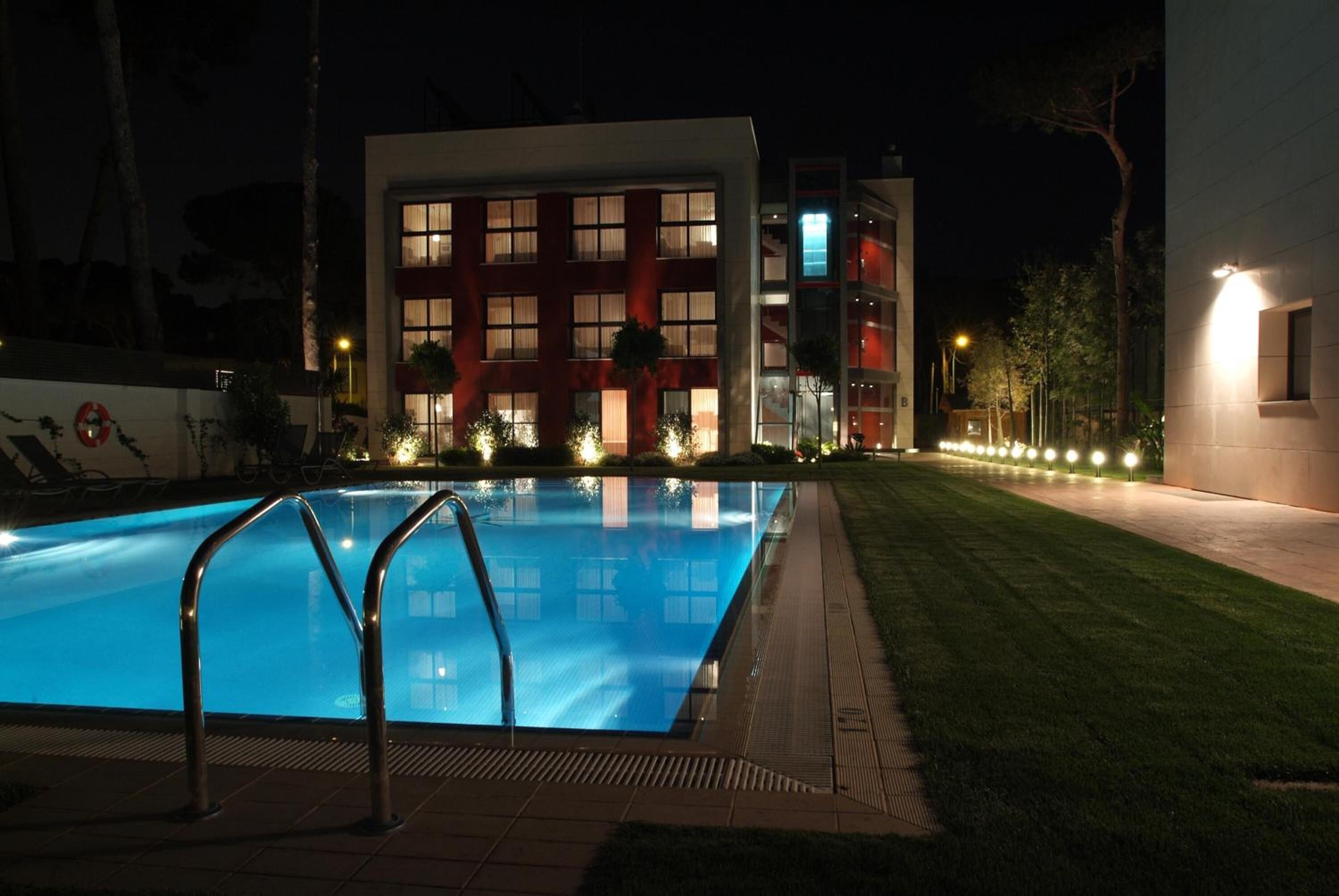 A swimming pool with metal railings, surrounded by a well-lit lawn and a building with multiple windows and balconies.