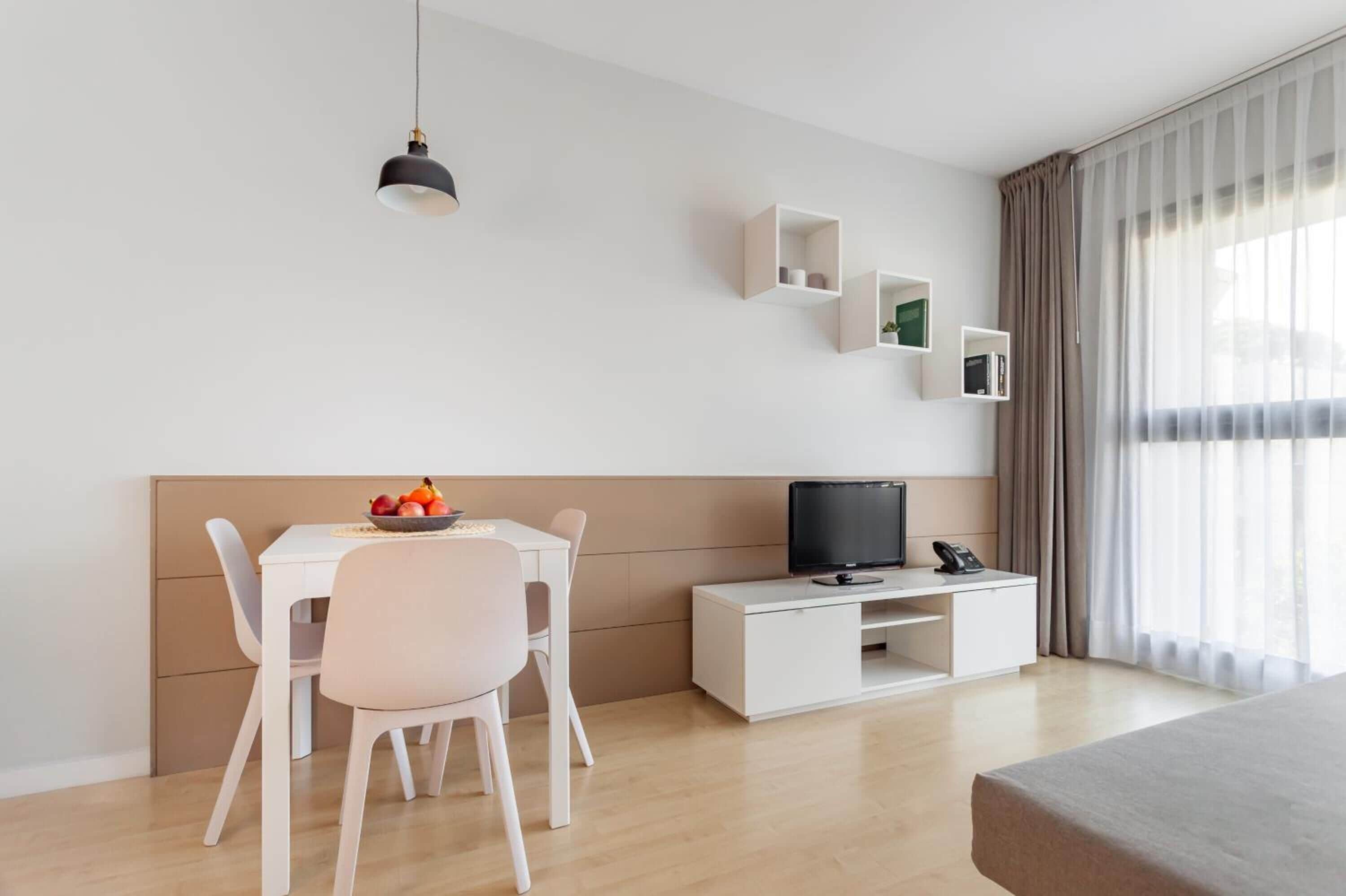 A modern hotel room with a bed, a desk with a computer, a chair, a small table with fruit, and a TV.