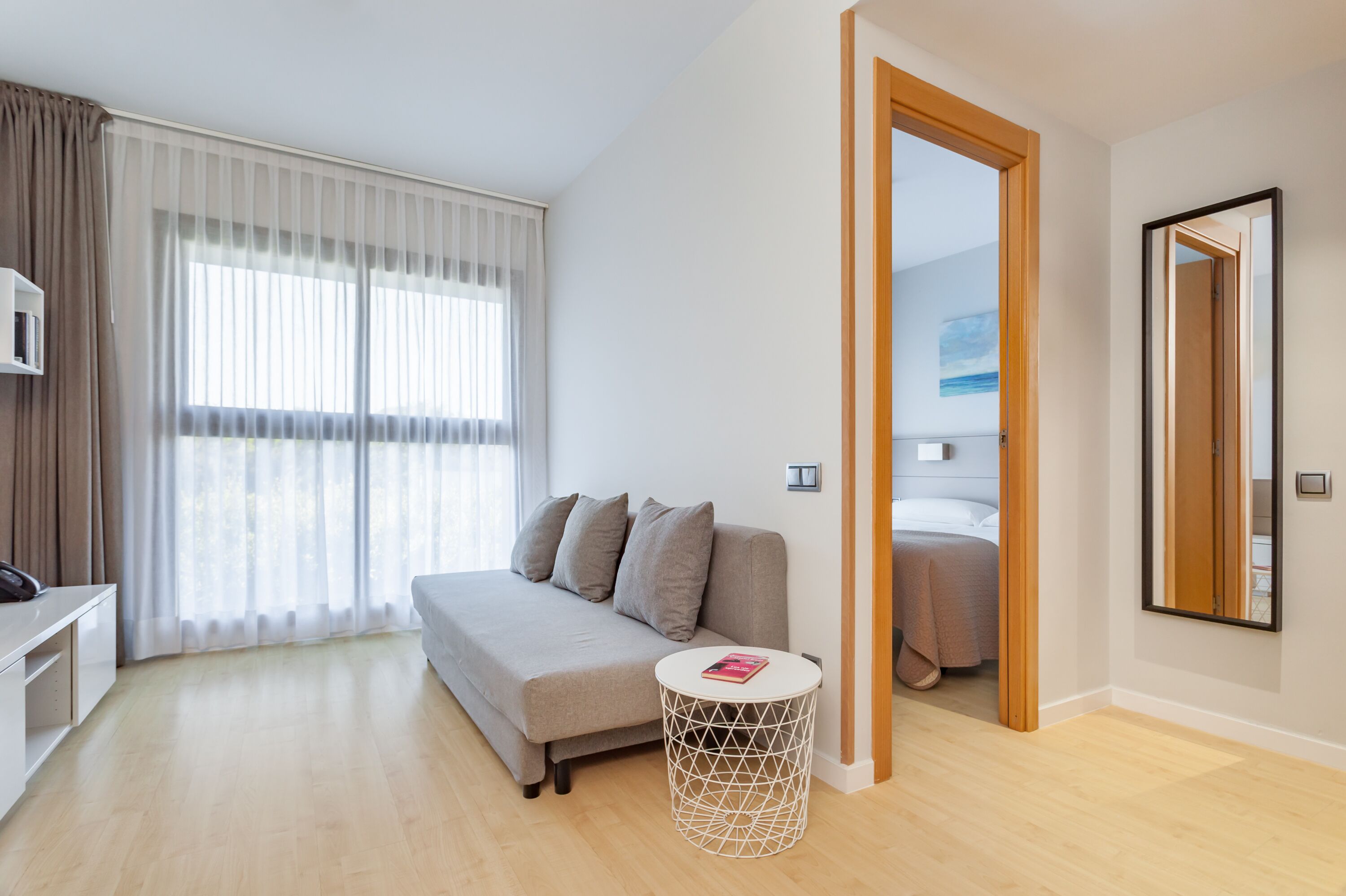 Una habitación moderna y bien iluminada con un sofá, una pequeña mesa redonda y una cama al fondo.