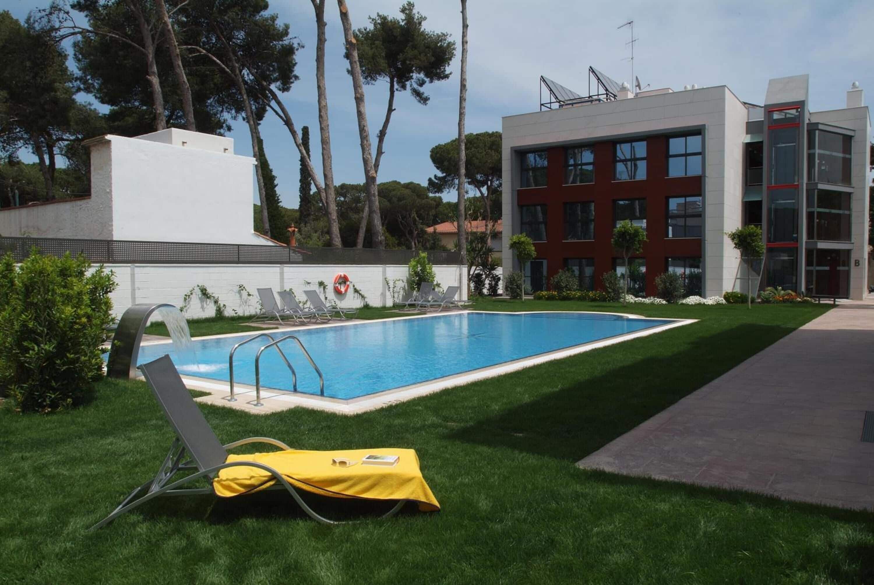En swimmingpool med liggestole og et gult håndklæde, omgivet af en velholdt græsplæne og moderne bygninger.
