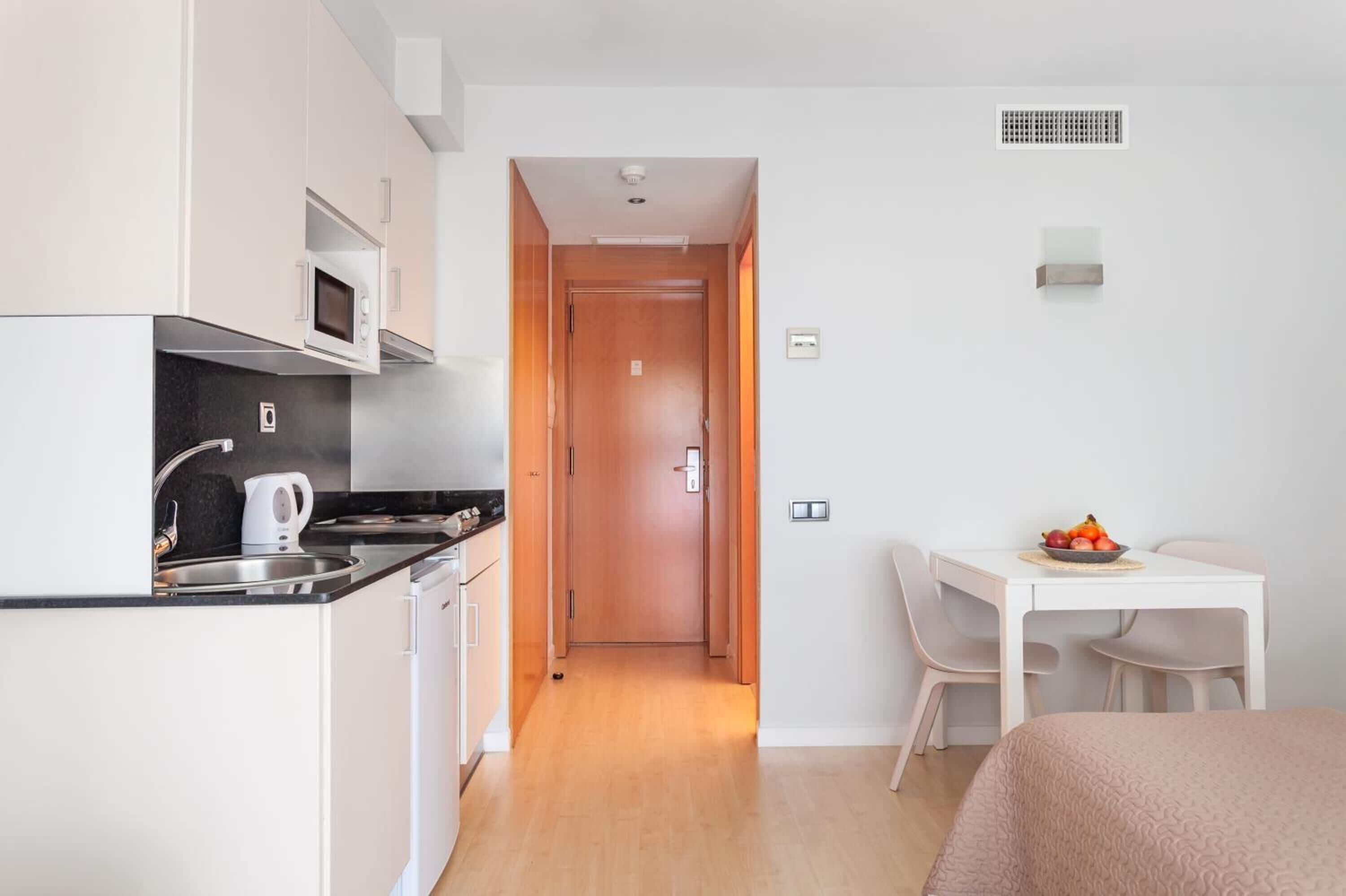 A compact kitchen with white cabinets, a microwave, and a kettle. A wooden door leads to a dining area with a table and chairs.