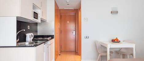 Een kleine keuken met witte kastjes, een magnetron en een waterkoker. Een houten deur leidt naar een eethoek met een tafel en stoelen.
