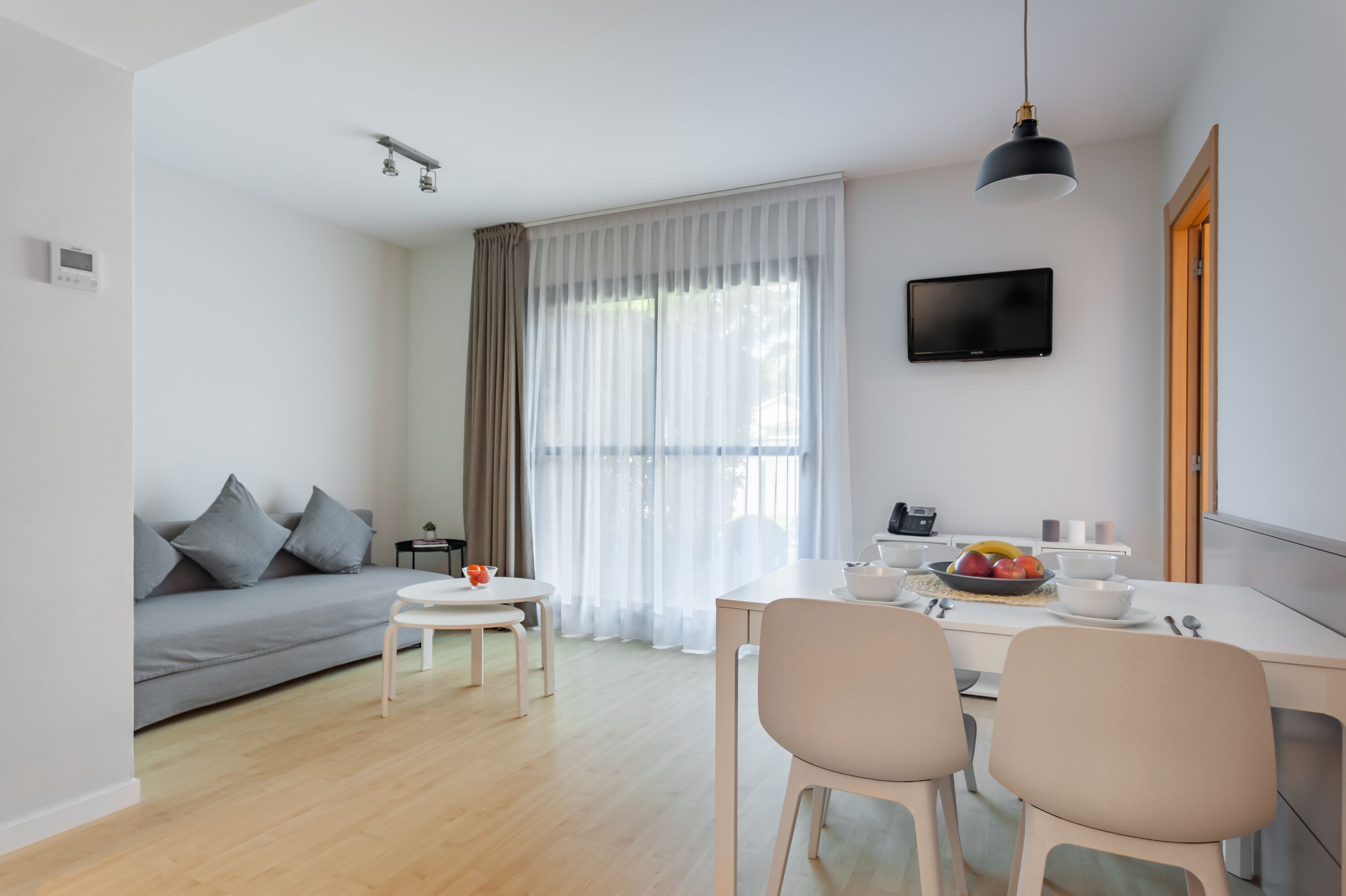 Un salon moderne avec un canapé gris, une table à manger avec des chaises, un téléviseur fixé au mur et une grande fenêtre ornée de rideaux légers.