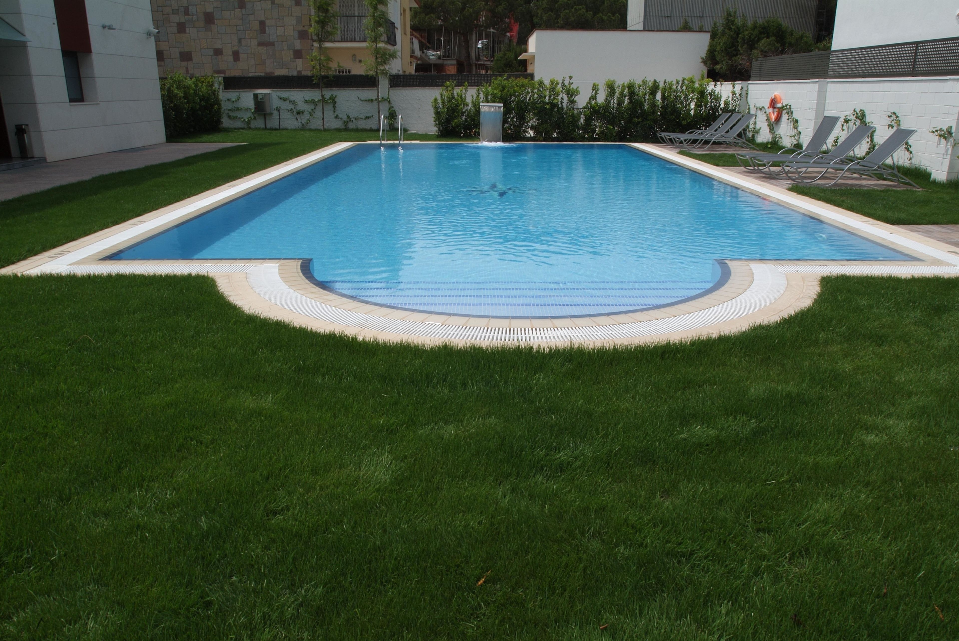Une piscine rectangulaire bordée de carreaux, entourée d’une pelouse bien entretenue et de quelques chaises longues.