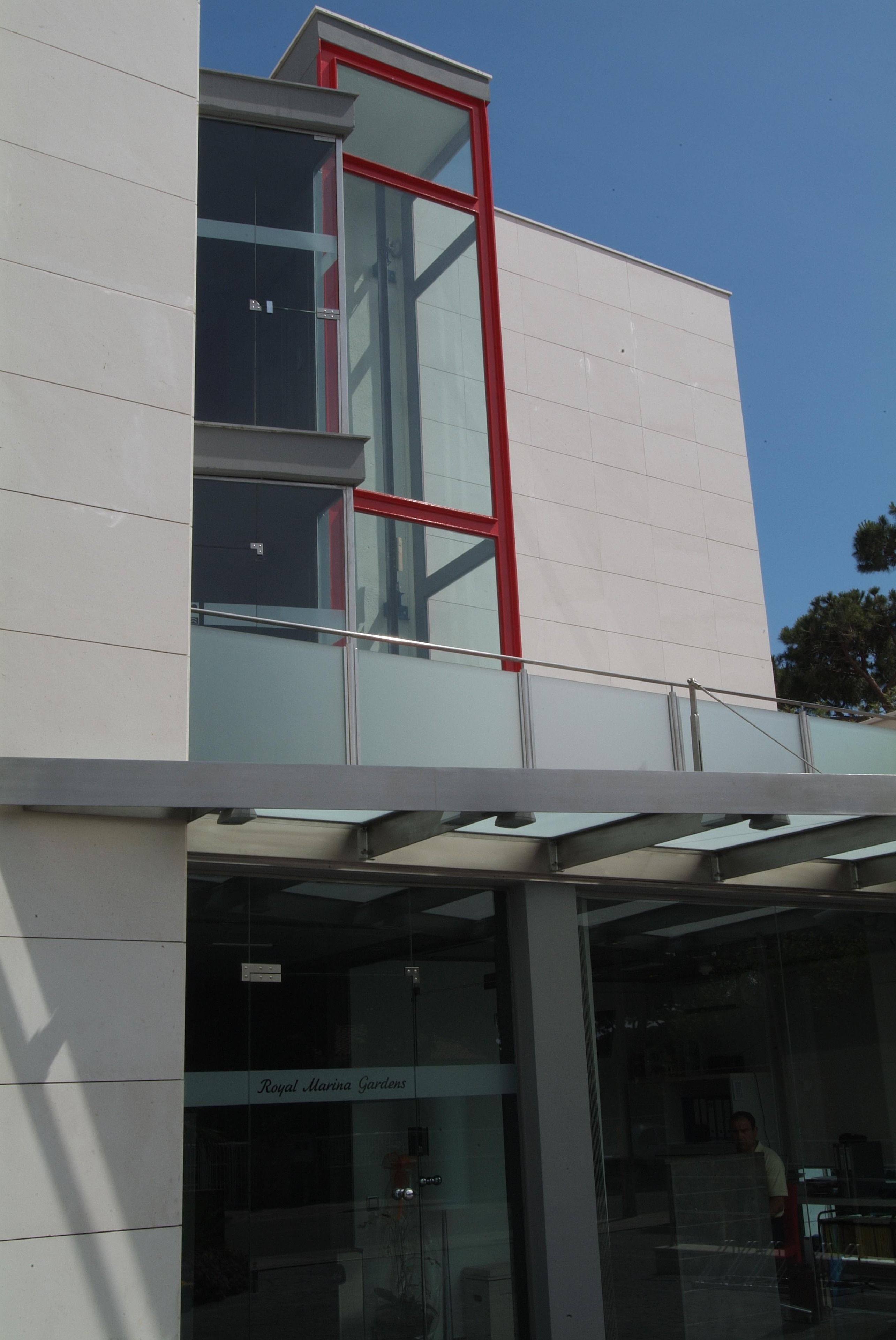 A modern building with a glass facade, a red frame around the windows, and a sign reading "Royal Marina Gardens".