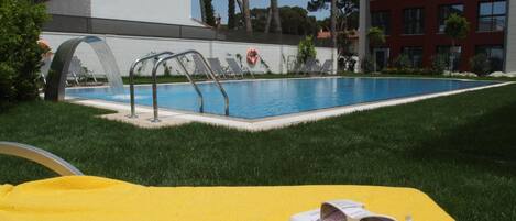 Une piscine agrémentée d’un élément décoratif aquatique, entourée d’une pelouse bien entretenue et d’un bâtiment à la façade rouge.