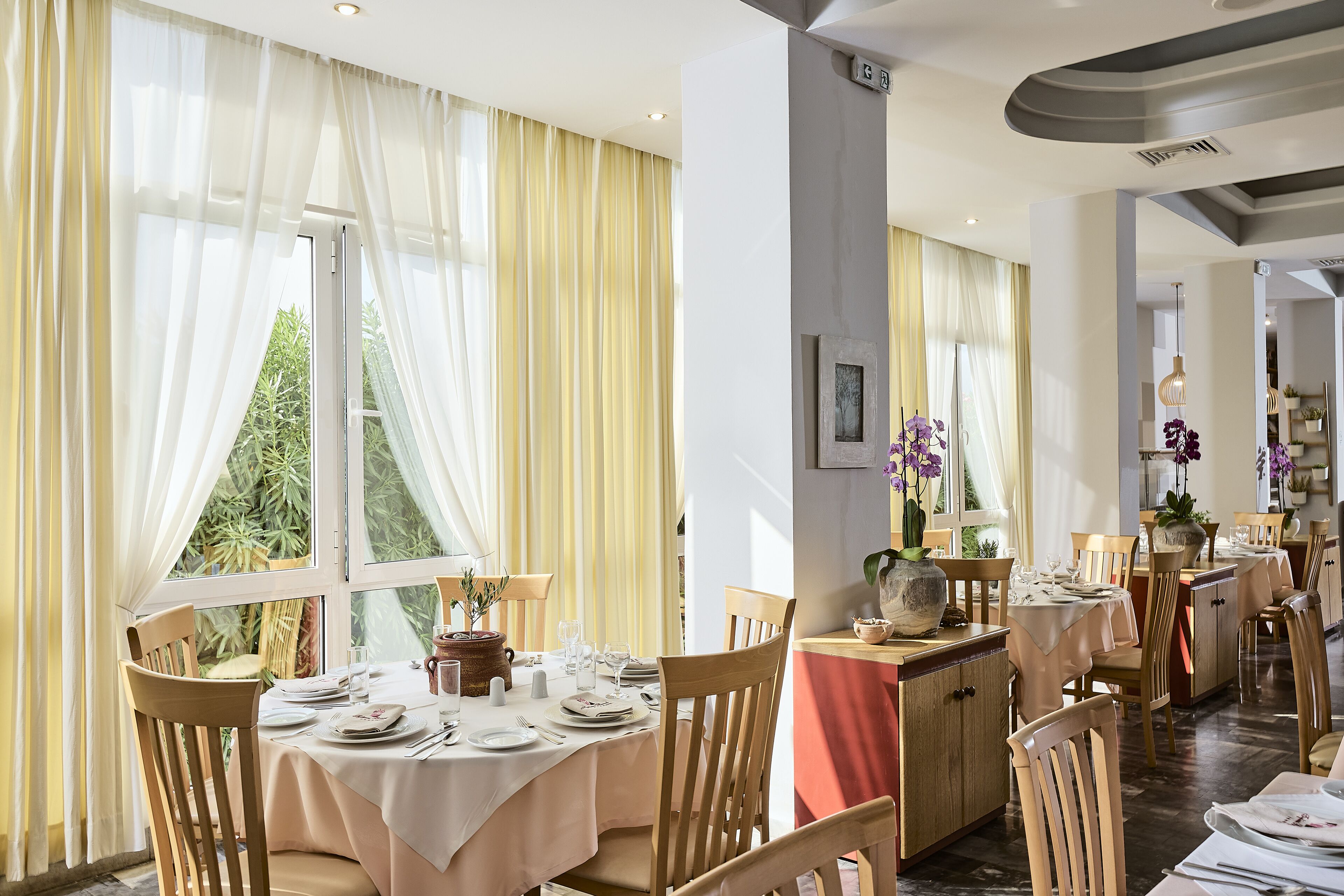 Un restaurante con mesas dispuestas para comer, ventanas amplias con cortinas y vistas al exterior con vegetación.