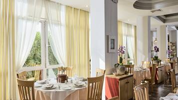 Un restaurante con mesas dispuestas para comer, ventanas amplias con cortinas y vistas al exterior con vegetación.